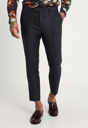 Slim-fit donkergrijze broek gedragen met bordeauxrode loafers met kwastjes, bloes zichtbaar aan de taille en getatoeëerde handen in de zakken.