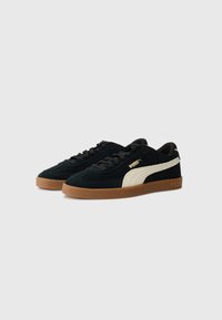 Black suede sneakers with a cream side stripe, featuring a brown rubber sole and black laces. Branding on the side and heel.