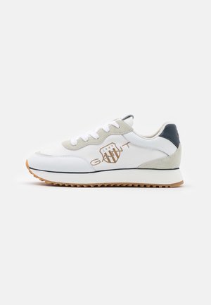 Zapatilla blanca y gris claro con cordones blancos, suela de goma texturizada, parche negro en el talón y logo marrón "GANT 1949" en el lateral.