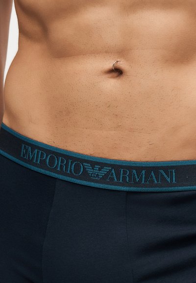 Κοντινό ενός γυμνού κοιλιακού με ασημένιο δαχτυλίδι ομφαλού και μαύρη μέση εσωρούχων Emporio Armani.