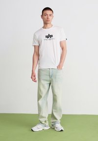 Hvid bomulds t-shirt med sort logo, parret med lysegrønne denimjeans med et falmet, tie-dye mønster og hvide sportssko.