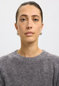 Pelēkā adītā džemperī ar apaļu kakla izgriezumu un strukturētu audumu. Modeles ausīs ir sudraba gredzenveida auskari, ar gludu ādas toni un dabiskiem grima akcentiem.