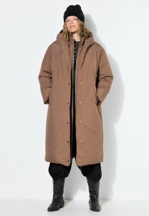 Una mujer luce un abrigo largo marrón con capucha, un gorro negro de punto, pantalones negros holgados y botas negras altas sobre un fondo blanco liso.