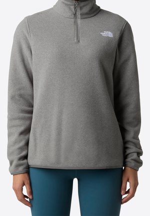 Osoba ubrana w szarą bluzę polarową z czwartym suwakiem z logo The North Face na piersi oraz turkusowe legginsy, widoczna od szyi do bioder.