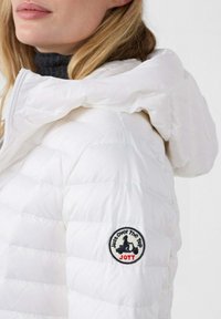 Giacca imbottita bianca con cappuccio, caratterizzata da una texture trapuntata e una toppa del logo rotonda sulla manica. Il tessuto appare lucido e leggero.