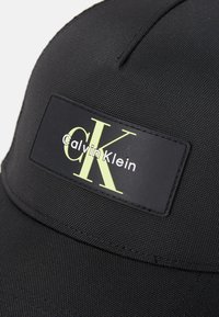 Sort kasket med struktureret stof, en buet skygge og et rektangulært logo-patch med "Calvin Klein" og "CK" i kontrasterende gulgrøn.