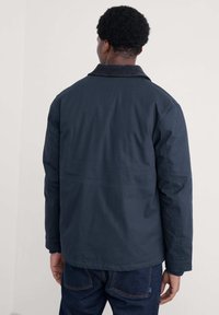 Jachetă ușoară, de culoare bleu închis, cu guler plat, mâneci lungi și o suprafață texturată, prezentată din spate, asortată cu blugi din denim închis.