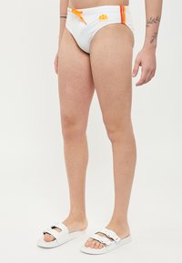 Slip de bain blanc avec des accents orange et un cordon de serrage ; présente un logo à l'avant. Associé à des sandales blanches à glissière avec des boucles doubles.