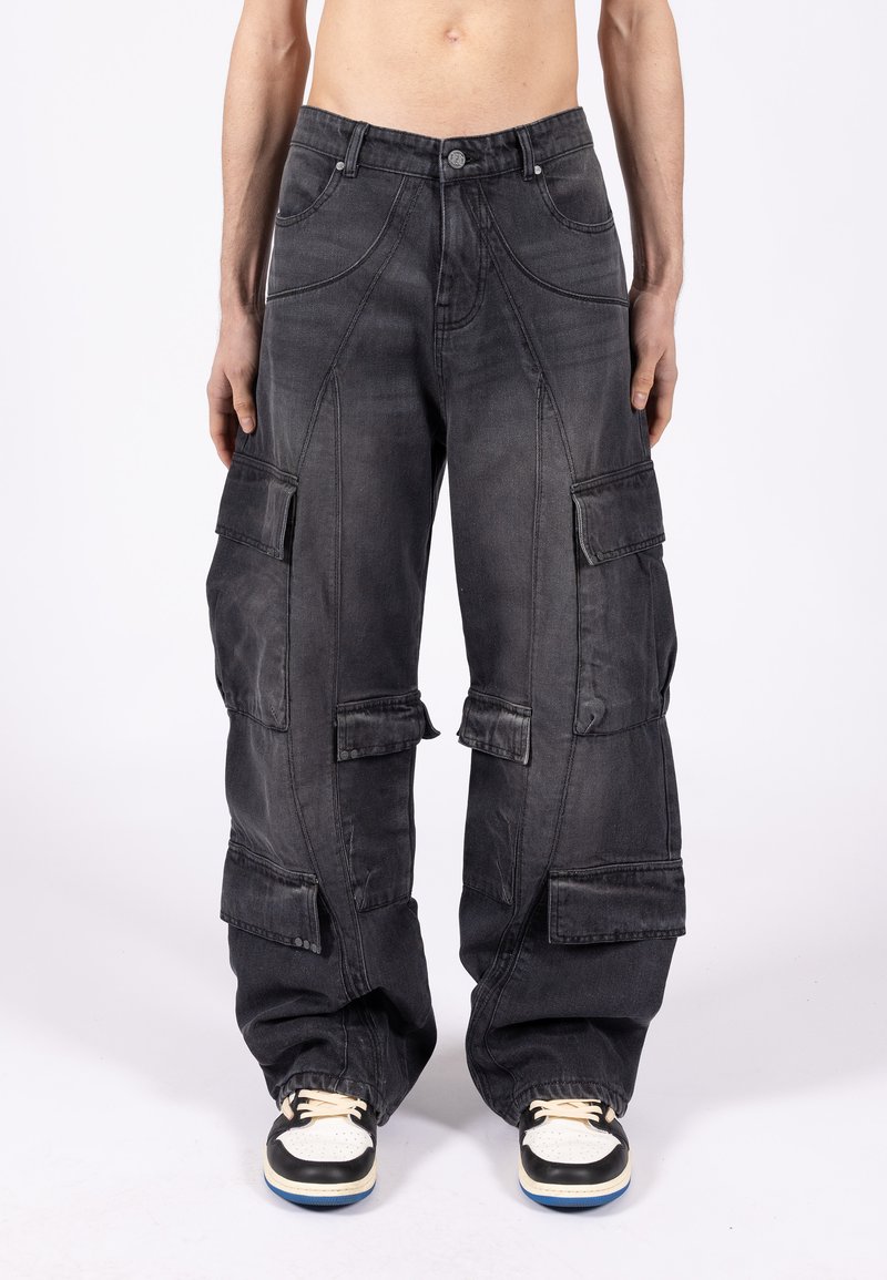 Mężczyzna w luźnych czarnych spodniach cargo z wieloma kieszeniami na klapki oraz w czarno-białych trampkach, stojący na tle jednolitego białego tła.