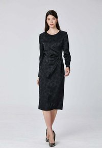 Robe noire à manches longues avec une texture florale, silhouette ajustée et détail enveloppant à la taille. Associée à des talons aiguilles noirs.