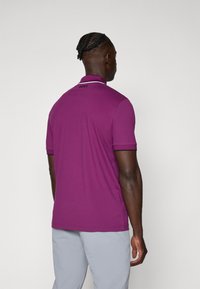 Vijolična polo majica s kratkimi rokavi, kontrastnim ovratnikom in logotipom na hrbtni strani. Material izgleda gladek z oprijetim dizajnom.