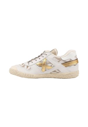 Zapatilla blanca con suela beige, detalles metálicos dorados que incluyen una gran X en el lateral y cordones marrón claro, mostrada desde el lado exterior.