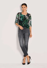 Mehrfarbiges Oberteil mit grünem und schwarzem Muster, Dreiviertelärmel, kombiniert mit engen grauen Jeans und schwarzen High Heels.