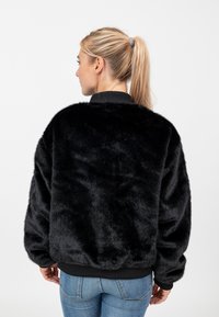 Giacca in finta pelliccia nera con vestibilità ampia, polsini neri a costine e scollatura rotonda. La texture è morbida e pelosa, adatta per la stagione più fresca.