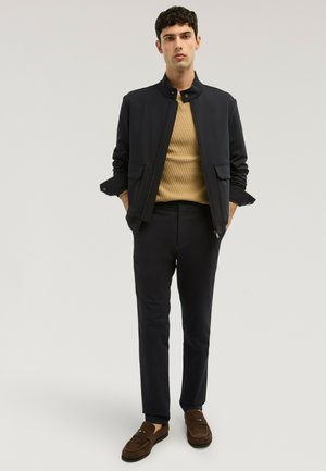 Junger Mann trägt schwarze Jacke, beige strukturierten Pullover, schwarze Hose und braune Loafers, steht mit den Händen in den Taschen vor einem einfarbigen Hintergrund.