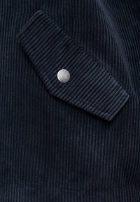 Κοντινό πλάνο υφάσματος κοτλέ σε ναυτικό μπλε χρώμα με ριβδωτή υφή και πτερύγιο ασφαλισμένο με ένα μόνο ασημένιο μεταλλικό κούμπωμα τύπου snap.