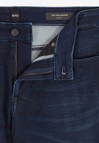 Jeans slim fit blu scuro con zip, bottone in metallo e cinturino interno in contrasto. Texture liscia con una leggera scoloritura.