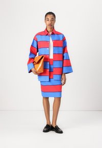 Blau-rote gestreifte Jacke mit Knöpfen, getragen über einem weißen Oberteil und einem passenden Rock. Hält eine orangefarbene Umschlagtasche und trägt schwarze Slipper.