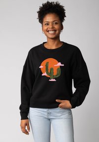Zwarte sweatshirt met een cactusontwerp, roze wolken en een levendige oranje zon. Zacht materiaal met een ontspannen pasvorm en geribbelde manchetten.