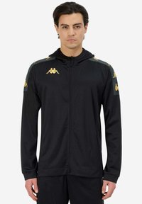 Giacca sportiva nera con zip e cappuccio, caratterizzata da dettagli dorati del logo Kappa sulle spalle e sulle maniche, realizzata in tessuto liscio con una vestibilità slim.
