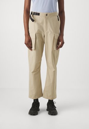 Persoon draagt beige cargobroek met voorvakken met klep, zwarte riem, zwarte sokken en zwarte stevige sneakers, staand tegen een effen achtergrond.