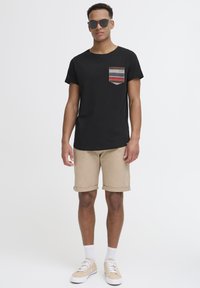 Svart t-skjorte med korte ermer og mønstrete lomme, beige shorts, hvite sokker og lyse sneakers. Solbriller fullfører det avslappede utseendet.