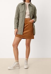 Chaqueta acolchada oliva con dos bolsillos en el pecho, top de canalé beige y falda de pana marrón. Zapatillas altas blancas con detalles texturizados.