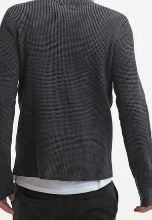 Man draagt een donkergrijze gebreide trui met textuur over een wit overhemd, gezien van achteren tegen een effen achtergrond.