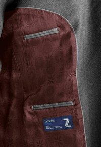 Grijze colbert interieur met een diep bordeauxrode voering met een subtiel patroon, twee zakken en een blauw stoffen label met verzorging-instructies.