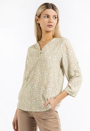 Jonge vrouw draagt een beige bloemenblouse met lange mouwen en een V-hals, poseert met één hand in de zak en kijkt naar opzij.