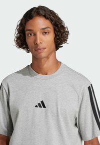 Sivá bavlnená tričko s okrúhlym výstrihom, krátkymi rukávmi a čiernymi pruhmi na pleciach. Na prednej strane sa nachádza čierne logo Adidas.