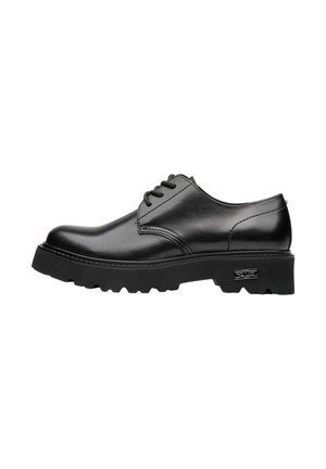 Scarpa nera in pelle con lacci, suola spessa scanalata e tacco basso, vista laterale su sfondo bianco.