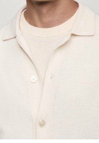 Beige knitted cardigan with a collar, featuring two buttons and layered over a matching beige sweater. Soft texture and subtle design.