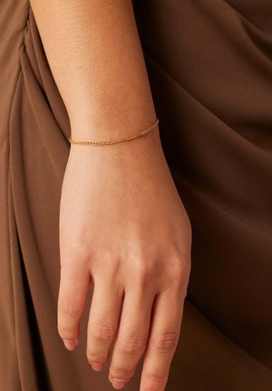 Gold chain bracelet on a wrist, featuring a delicate design with fine links. The hand rests against a brown fabric background.