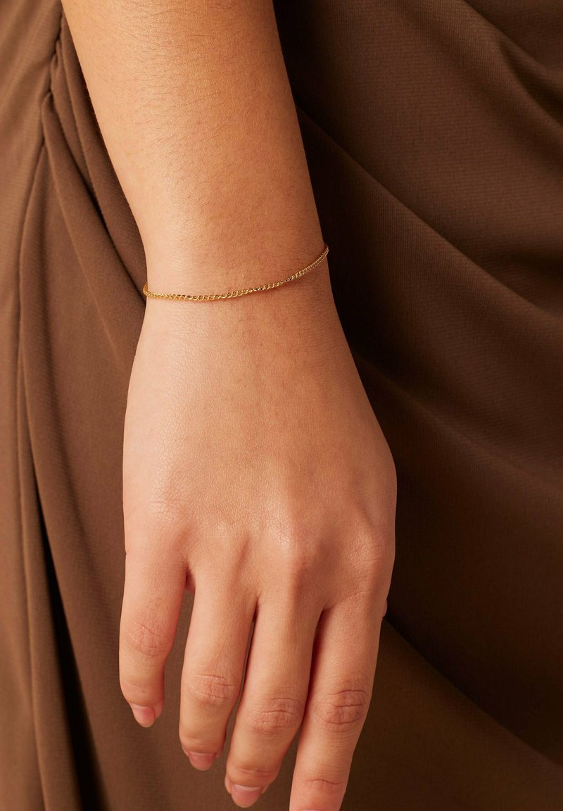 Bracelet en chaîne dorée au poignet, présentant un design délicat avec des maillons fins. La main repose sur un fond en tissu marron.