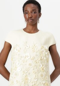 White textured dress with short sleeves, featuring intricate floral embellishments in pale yellow and white, embellished with sequins.