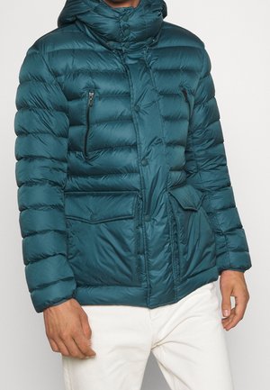 Mann trägt eine türkisfarbene Steppjacke mit Kapuze, Reißverschluss und Knopfverschlüssen, mehreren Vordertaschen, kombiniert mit weißer Hose.