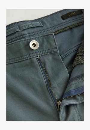 Housut, joissa on kapea leikkaus, valmistettu teksturoidusta vihreästä kankaasta, varustettu metallisella nappilukolla, vetoketjulla ja erottuvalla sisävyötärön kuviolla.