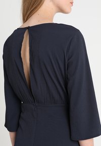 Tmavě modré šaty s texturovanou tkaninou, s kulatým výstřihem vzadu, 3/4 rukávy a jemným stahovaným pasem s uzávěrem na zadní straně.