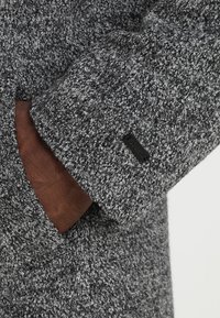 Grijze textuur jas gemaakt van een wol-mix stof. Bevat een bruine leren manchettenaccent en een klein zwart merkje op de mouw.