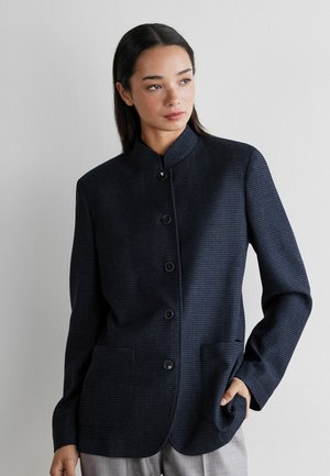 Kobieta o prostych, ciemnych włosach, ubrana w zapinany na guziki ciemnoniebieski, teksturowany żakiet ze stójką, z ręką w kieszeni, skierowana nieco w prawo.