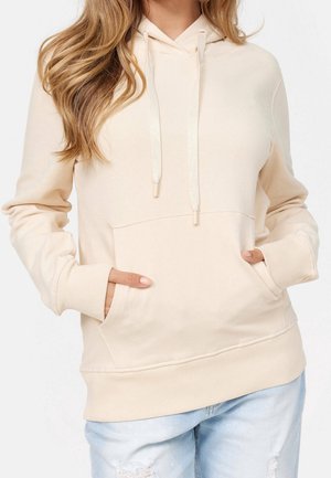 Un hoodie beige en tissu doux, avec une poche avant, des cordons de serrage et des poignets côtelés, associé à un jean déchiré de couleur claire.