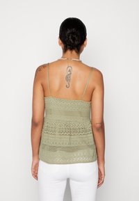 Krajkový top na ramínka v tlumené zelené barvě s tenkými popruhy. Obsahuje složité vzory a peplum střih. Nošený s bílými kalhotami.