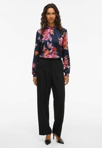 Blusa floreale in blu navy con motivi viola, arancioni e rosa; scollatura alta; abbinata a pantaloni neri su misura e tacchi neri a punta.