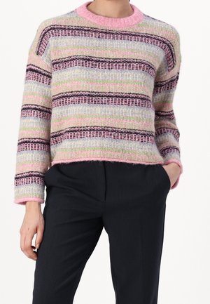 Personne portant un pull tricoté à rayures pastel avec des tons de rose, violet, vert et bleu, assorti à un pantalon noir, la main dans la poche.