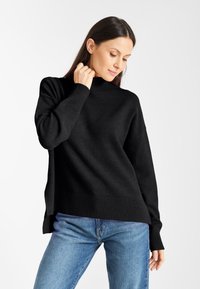 Schwarzer Strickpullover mit hohem Kragen und gerippten Bündchen. Das Design zeichnet sich durch eine lockere Passform und seitliche Schlitze aus, kombiniert mit blauen Jeans.