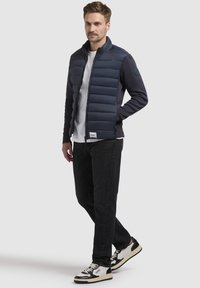 Mørkeblå puffervest med quiltet front og strikkede ærmer. Parret med en hvid T-shirt, sorte jeans og sorte/hvide sneakers. Glat tekstur.