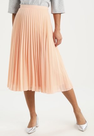 Woman wearing a peach pleated midi skirt and silver block-heeled shoes, standing against a plain white background.