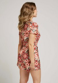 Salopette à motifs floraux en rouge, noir et crème ; manches courtes, taille cintrée et coupe décontractée, fabriquée en tissu léger.