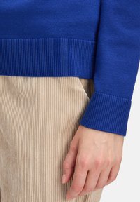 Kék kötött pulcsi bordás mandzsettával, bézs kordbársony nadrággal, függőleges csíkokkal és sima textúrával párosítva.